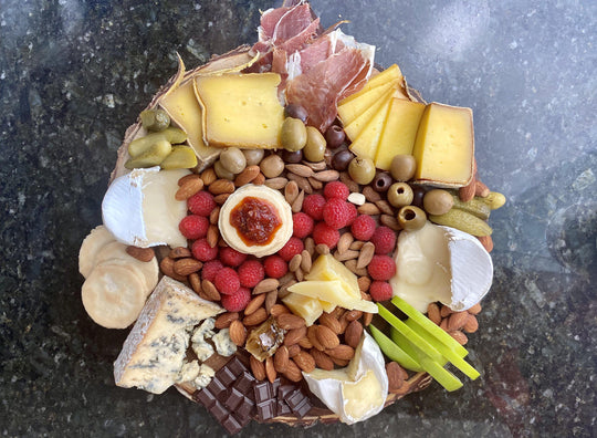 a round cheese and charcuterie board with nuts, fruit, chocolate, and olives