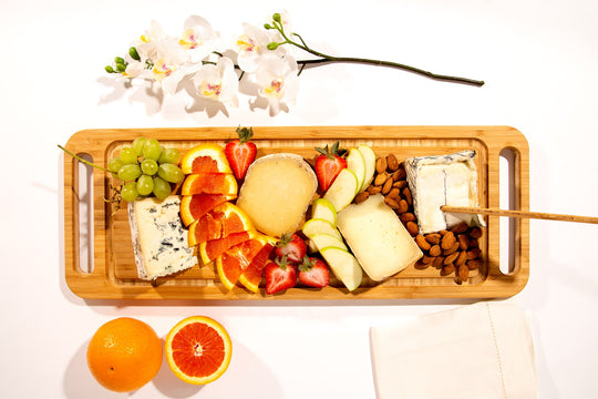overhead view of rectangular bamboo cheese board with hadles lade with fruit, cheese, and nuts with flowers and cut orange next to it