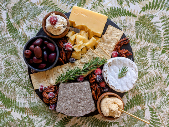 round slate cheese board with cheese, pate, olives, crackers, and nuts on fern tablecloth